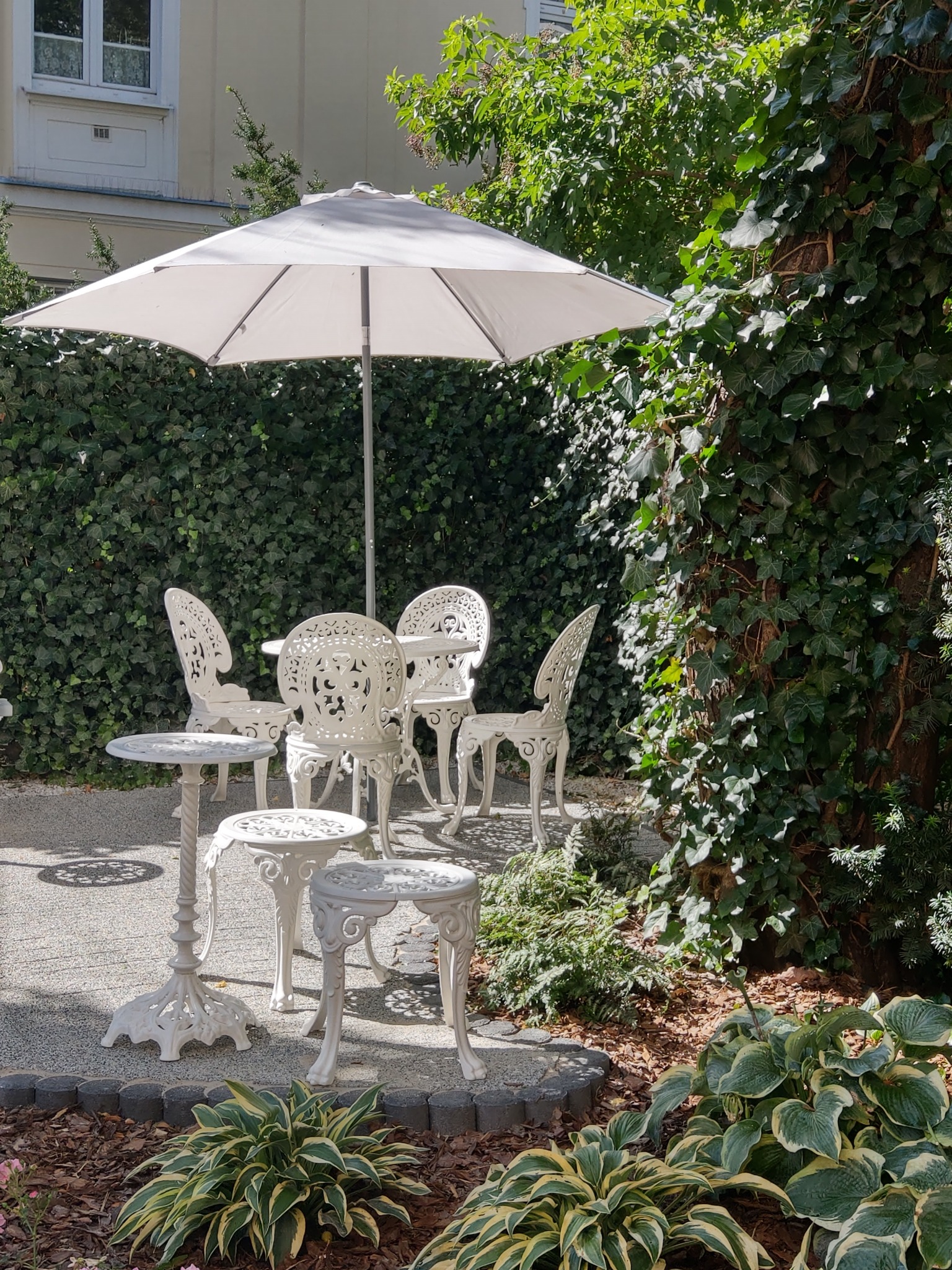 Biały, ażurowy zestaw mebli ogrodowych pod parasolem na tle ściany z bluszczu i roślinności.