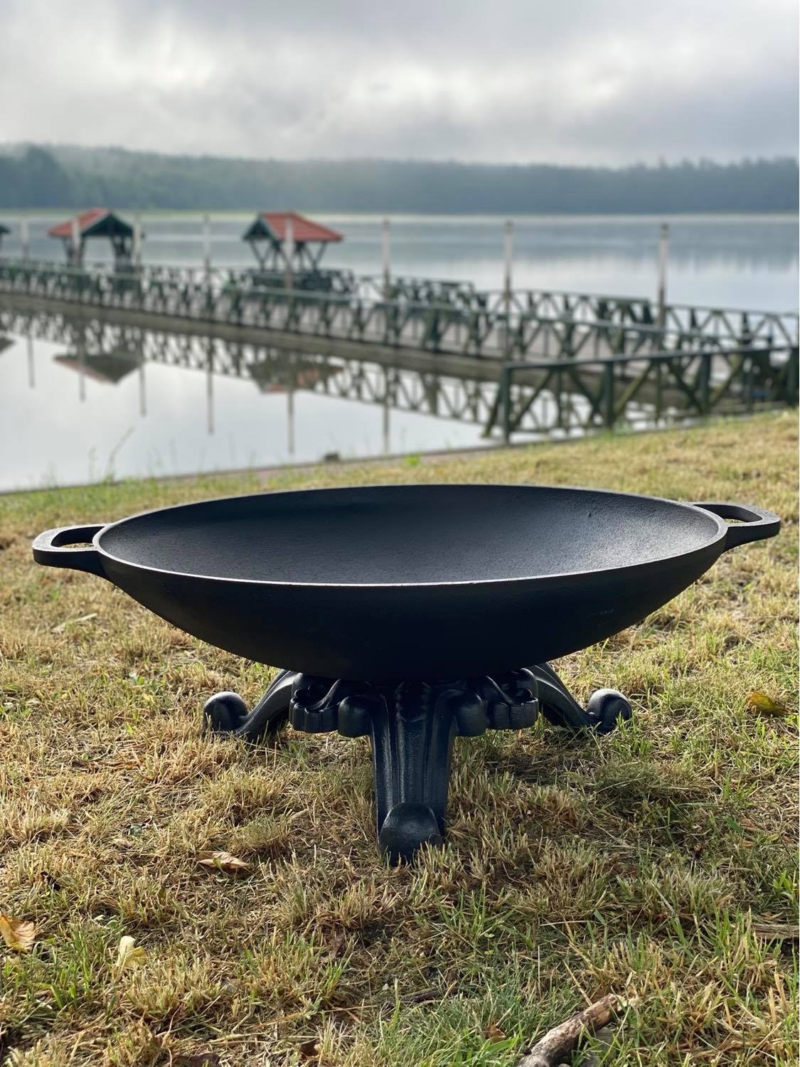 Czarny, metalowy palenisko ogrodowe w kształcie misy na ozdobnej podstawie, ustawione na trawie, z widokiem na jezioro i pomost w tle, pochmurny dzień.