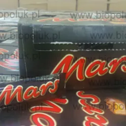 Karton zbiorczy batonów Mars, na tle innych kartonów, z pojedynczym batonem na wierzchu, widoczne logo firmy oraz adres strony internetowej na opakowaniach.