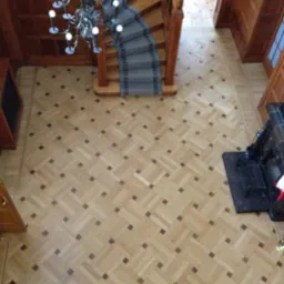 Wysoki kąt widzenia na parkiet z jasnego drewna ułożony we wzór jodełki z ciemnymi akcentami, widoczne schody z szarą wykładziną i żyrandol z białymi kulami