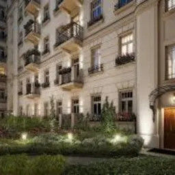 Elewacja kamienicy z zadbanym, oświetlonym ogrodem przed wejściem. Widoczne balkony z roślinami i ozdobne detale architektoniczne. Oświetlona brama wejściowa.