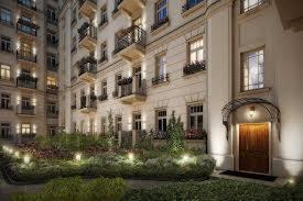 Elewacja kamienicy z zadbanym, oświetlonym ogrodem przed wejściem. Widoczne balkony z roślinami i ozdobne detale architektoniczne. Oświetlona brama wejściowa.