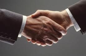 Uścisk dłoni dwóch mężczyzn w garniturach, symbolizujący zawarcie umowy lub partnerstwo biznesowe, z widocznym mankietem koszuli jednego z mężczyzn.