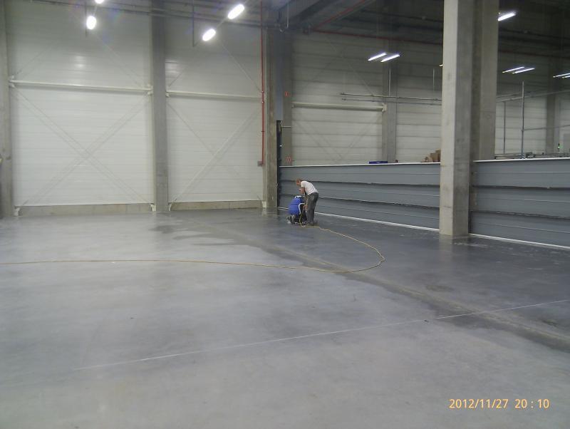 Pracownik w białej koszulce czyści posadzkę przemysłową w hali magazynowej za pomocą szorowarki z niebieskim zbiornikiem; widoczne ślady wilgoci po czyszczeniu.