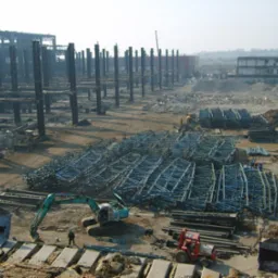 Rozległy teren po rozbiórce hali przemysłowej z pozostałościami konstrukcji, stertami gruzu i przygotowanymi elementami stalowymi; na pierwszym planie koparka i mały ciągnik.