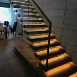 Nowoczesne schody z podświetlanymi stopniami, szklaną balustradą i metalową poręczą, na tle ściany z płyt imitujących beton.