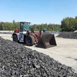 Ładowarka kołowa marki O&K z pełną łyżką ekogroszku na placu składowym, w tle hałdy węgla i zieleń drzew.