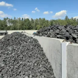 Dwa duże pryzmy węgla kamiennego oddzielone betonową ścianą na składzie opału, z widocznym samochodem ciężarowym i lasem w tle pod błękitnym niebem.