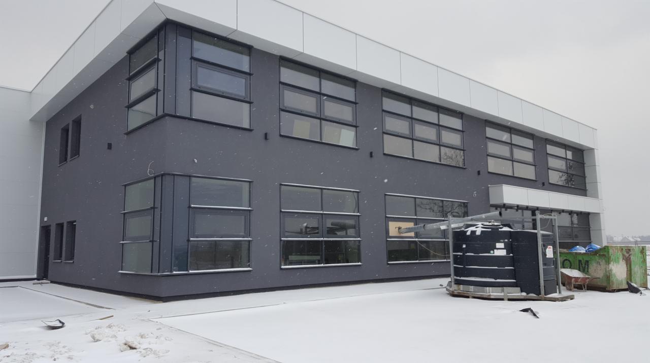 Nowoczesny budynek biurowy z szarymi ścianami i dużymi oknami, pokryty śniegiem podczas lekkich opadów.