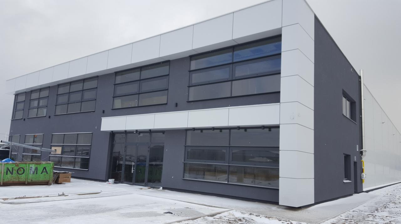 Nowoczesny budynek biurowy z szarymi ścianami i dużymi, prostokątnymi oknami z ciemnymi ramami, częściowo ośnieżony teren przed budynkiem.