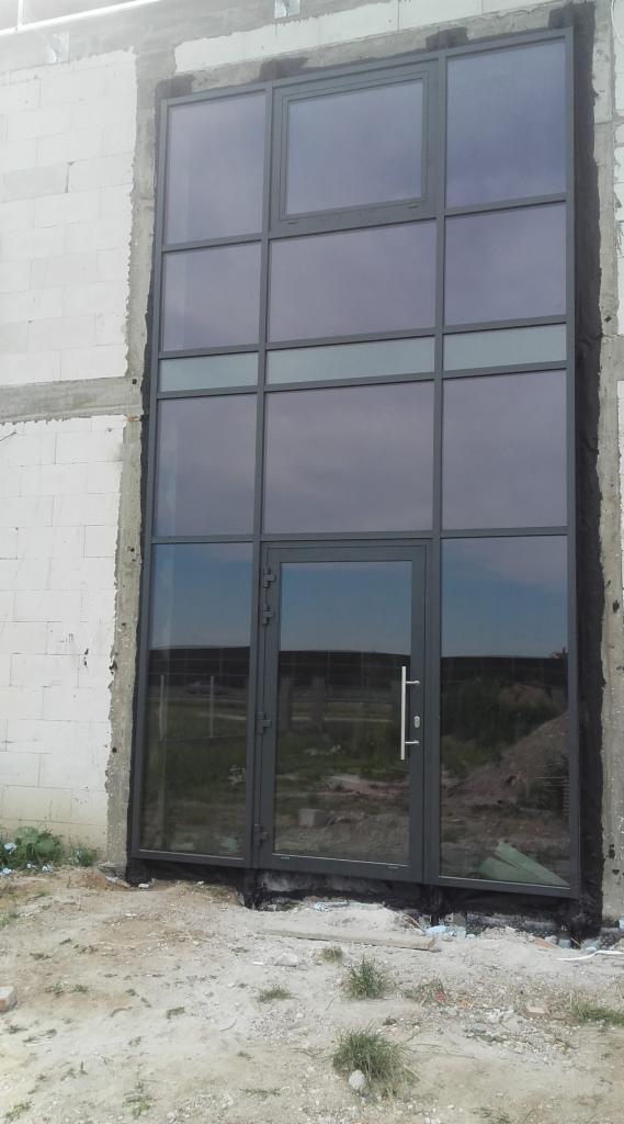 Wysoka, szara konstrukcja okienno-drzwiowa z wieloma szybami, w trakcie montażu w surowym budynku z białej cegły, widoczne niedokończone obrzeża i teren budowy.