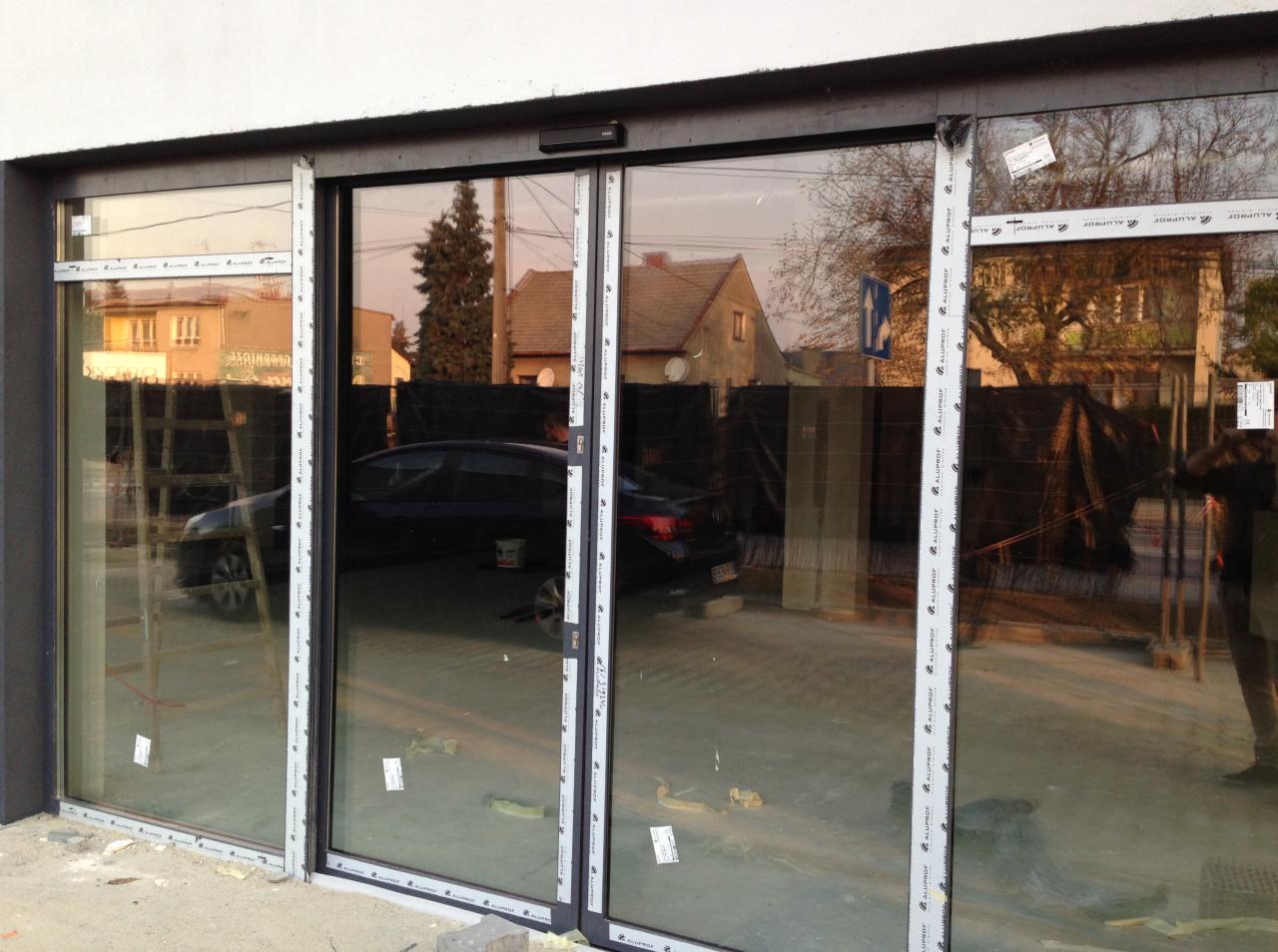 Zewnętrzny widok przeszklonej ściany z przesuwnymi drzwiami, czarne ramy z białymi listwami ochronnymi, widoczne odbicie otoczenia: budynki, samochód, drzewa, plac budowy.