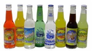 Rząd ośmiu szklanych butelek z kolorowymi napojami gazowanymi różnych marek, ustawionych na białym tle.