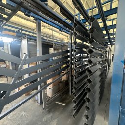 MAR-BET Ogrodzenia Czujak - Szare, metalowe profile ogrodzeniowe zawieszone na linii produkcyjnej w hali. Widoczne opakowania i regały w tle. Elementy przygotowane do montażu ogrodzenia.