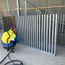 MAR-BET Ogrodzenia Czujak - Malowanie proszkowe metalowego panelu ogrodzeniowego w warsztacie. Pracownik w kombinezonie i masce ochronnej aplikuje farbę na pionowe elementy ogrodzenia.