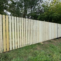 MAR-BET Ogrodzenia Czujak - Nowe ogrodzenie drewniane z zaokrąglonymi sztachetami, widoczne mocowania, świeże drewno, trawa w tle, częściowo zamontowane, zieleń drzew w tle.