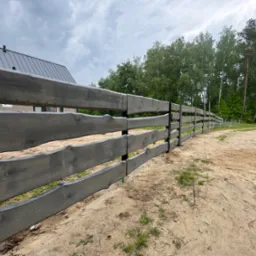 Długi, drewniany płot w kolorze szarości z surowym wykończeniem desek, otaczający posesję. W tle widoczne drzewa i fragment budynku z metalowym dachem.