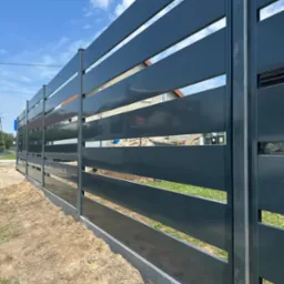 Nowoczesne, poziome ogrodzenie panelowe w kolorze grafitowym, z widocznymi prześwitami między panelami. W tle dom jednorodzinny pod błękitnym niebem.