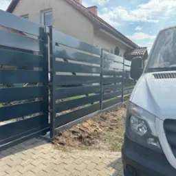 Nowoczesna, przesuwna brama panelowa w kolorze grafitowym, widoczna z perspektywy ulicy, z białym samochodem dostawczym po prawej stronie. Dom jednorodzinny w tle.