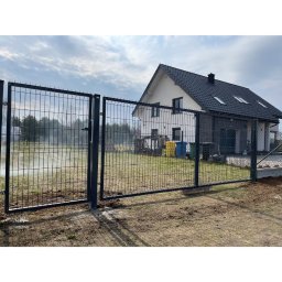MAR-BET Ogrodzenia Czujak - Nowoczesne ogrodzenie panelowe w kolorze grafitowym, z otwartą bramą wjazdową, na tle domu jednorodzinnego z ciemną dachówką i jasną elewacją. Widok z zewnątrz posesji.