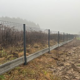 MAR-BET Ogrodzenia Czujak - Szary panelowy płot z podmurówką na zewnątrz, w tle zalesiony teren spowity mgłą. Widoczne słupki i siatka ogrodzeniowa. Teren wokół ogrodzenia jest nierówny i błotnisty.