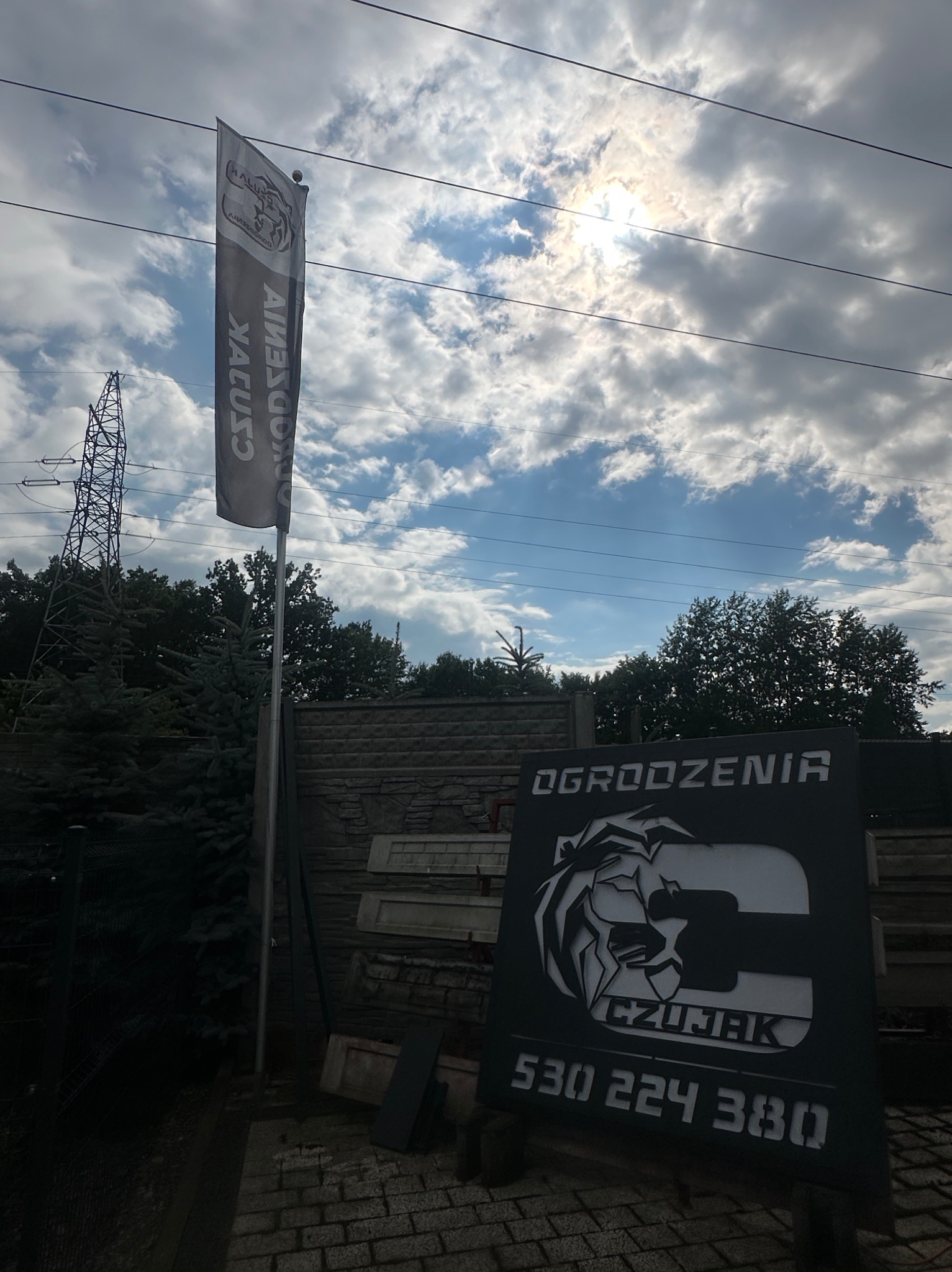 Tablica reklamowa firmy ogrodzeniowej 'Czułjak' z numerem telefonu na tle nieba i fragmentu ogrodzenia betonowego. Widoczny także baner firmy i słup wysokiego napięcia.