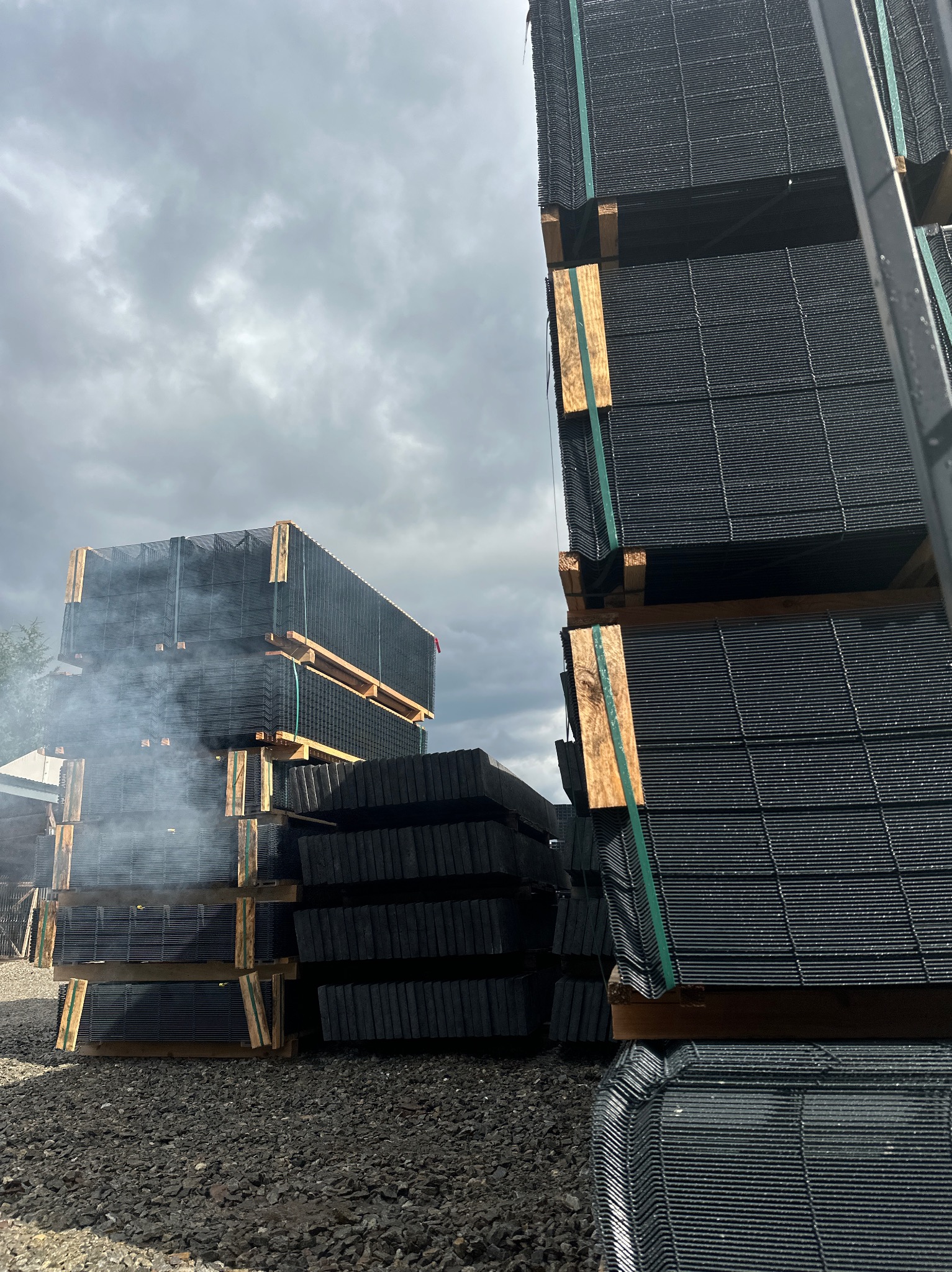 Stosy paneli ogrodzeniowych na paletach, zabezpieczonych taśmami, na żwirowym podłożu, z lekką mgłą w tle. Pochmurne niebo.