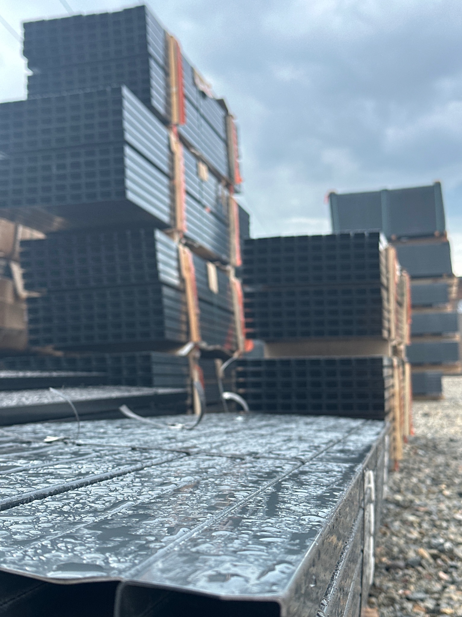 Stosy profili do ogrodzeń panelowych w Szczytnie, zabezpieczone taśmą. Widoczne krople wody na metalu, pochmurne niebo w tle. Materiał do montażu ogrodzenia.