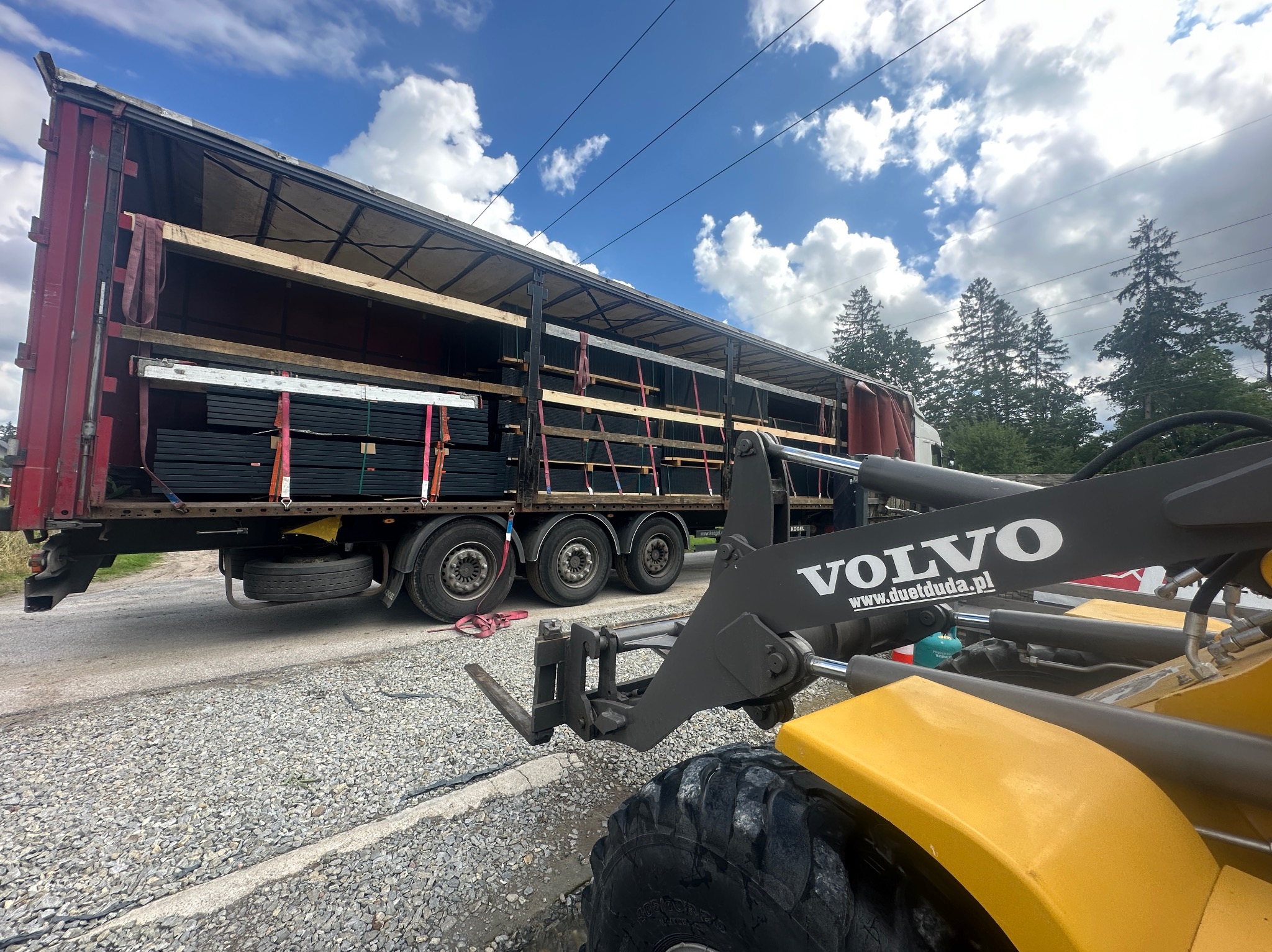 Rozładunek paneli ogrodzeniowych z ciężarówki za pomocą ładowarki Volvo na drodze. Widoczne niebo z chmurami i drzewa w tle.