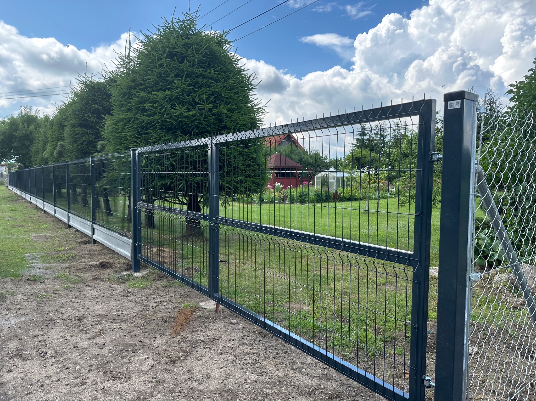 Nowoczesne ogrodzenie panelowe w kolorze grafitowym z otwartą bramą wjazdową. W tle widoczne drzewa iglaste i budynek mieszkalny.