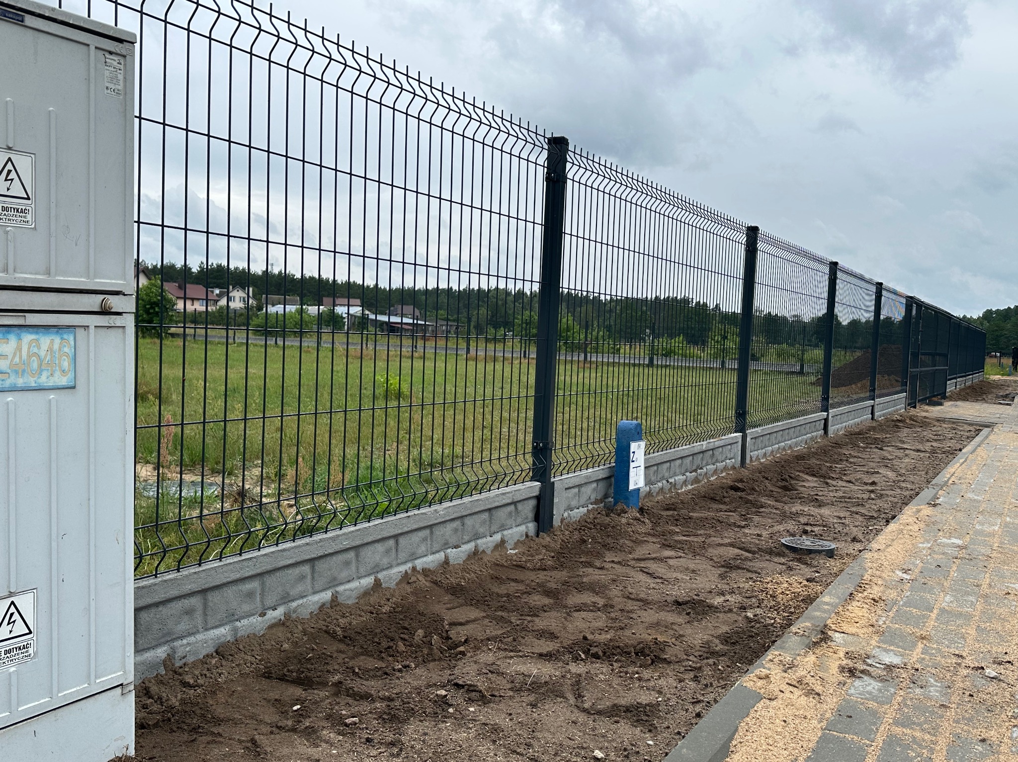 Nowoczesne ogrodzenie panelowe z podmurówką betonową, widok z boku, z elementami infrastruktury energetycznej w tle.