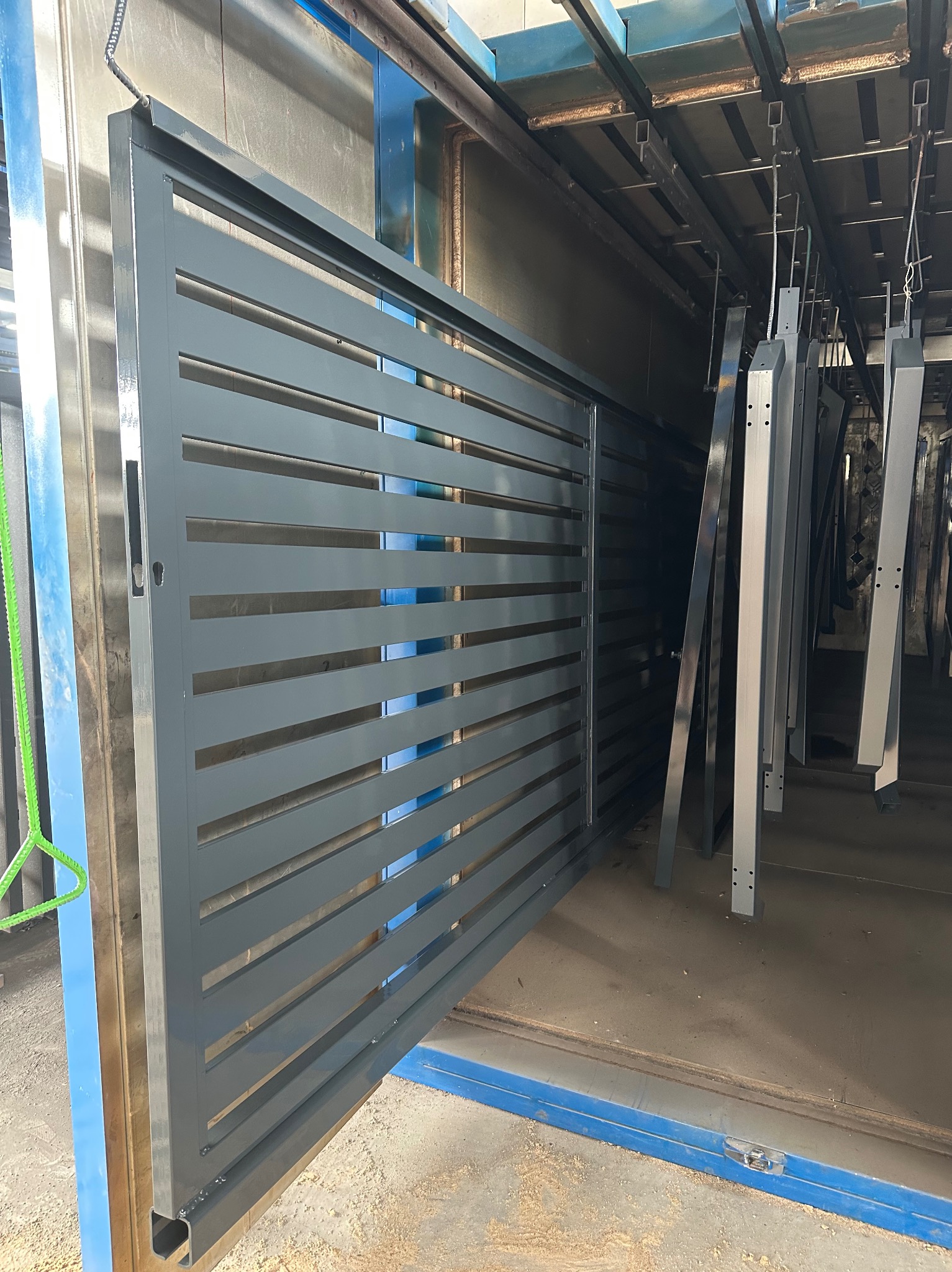 Szara, metalowa brama lamelowa w lakierni proszkowej, zawieszona na hakach, gotowa do malowania. Widoczne elementy konstrukcyjne i detale wykończenia.