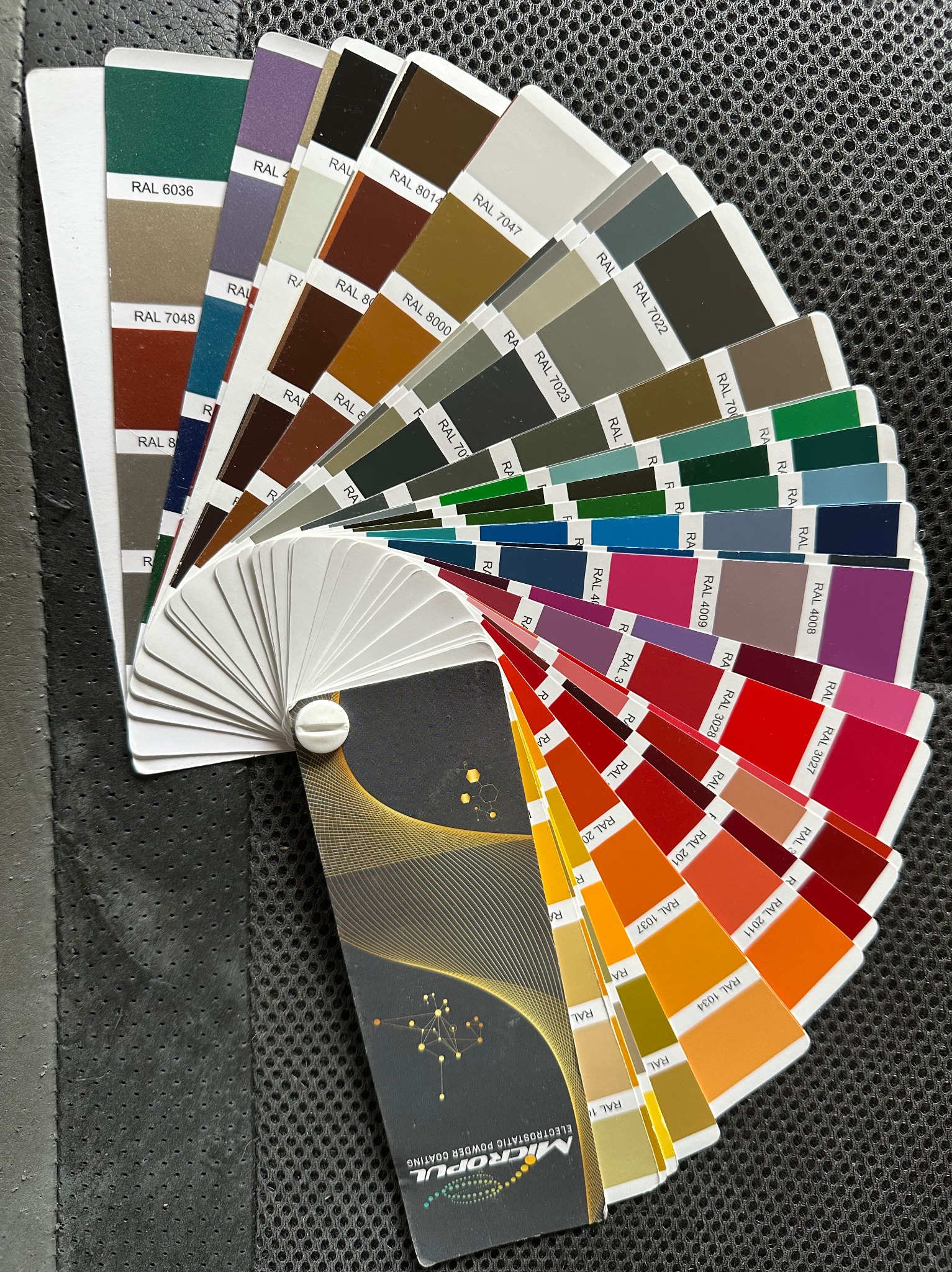 Paleta kolorów RAL Micropul z odcieniami farb proszkowych, ułożona wachlarzowo na ciemnym, perforowanym tle, prezentująca szeroki zakres barw od zieleni po czerwienie.