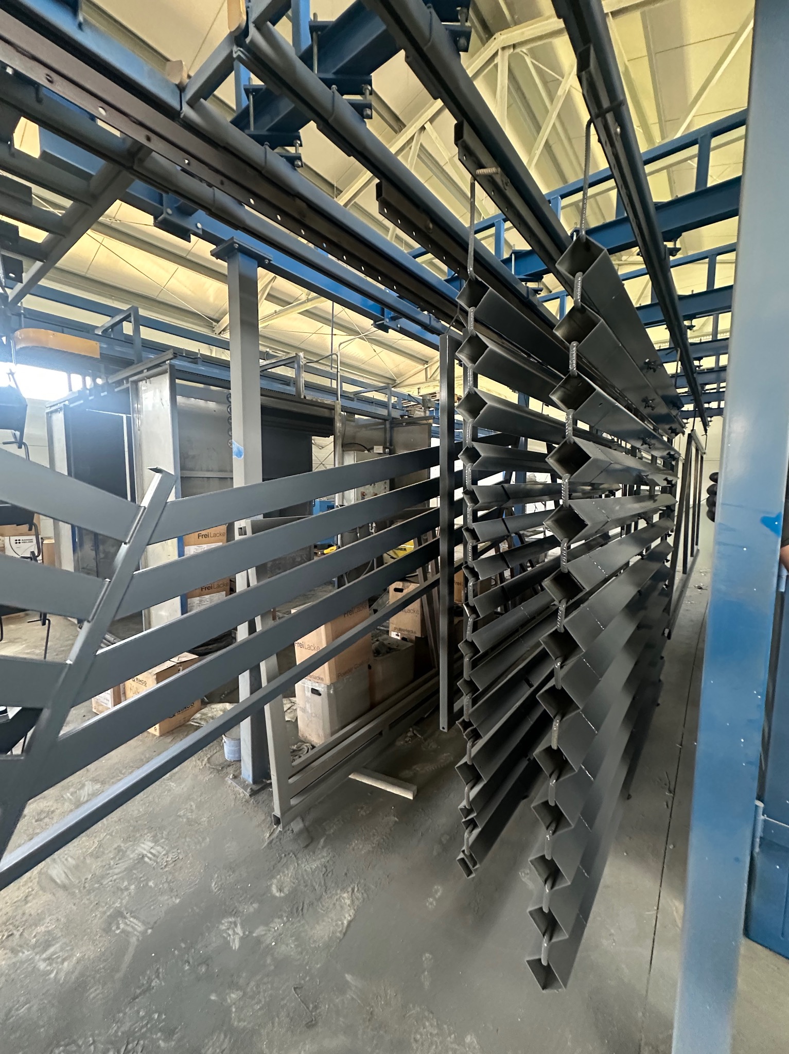 Szare, metalowe profile ogrodzeniowe zawieszone na linii produkcyjnej w hali. Widoczne opakowania i regały w tle. Elementy przygotowane do montażu ogrodzenia.