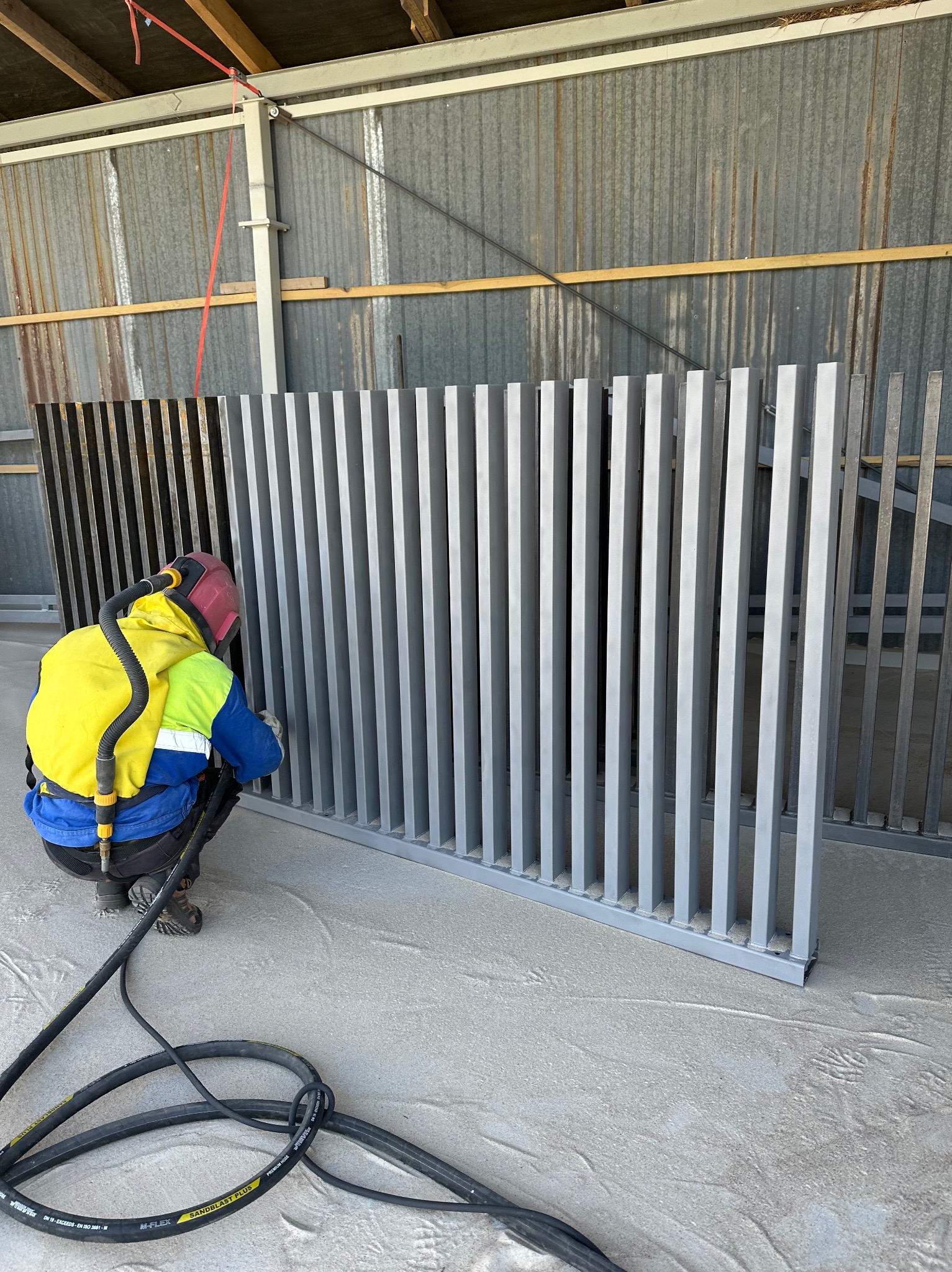 Malowanie proszkowe metalowego panelu ogrodzeniowego w warsztacie. Pracownik w kombinezonie i masce ochronnej aplikuje farbę na pionowe elementy ogrodzenia.