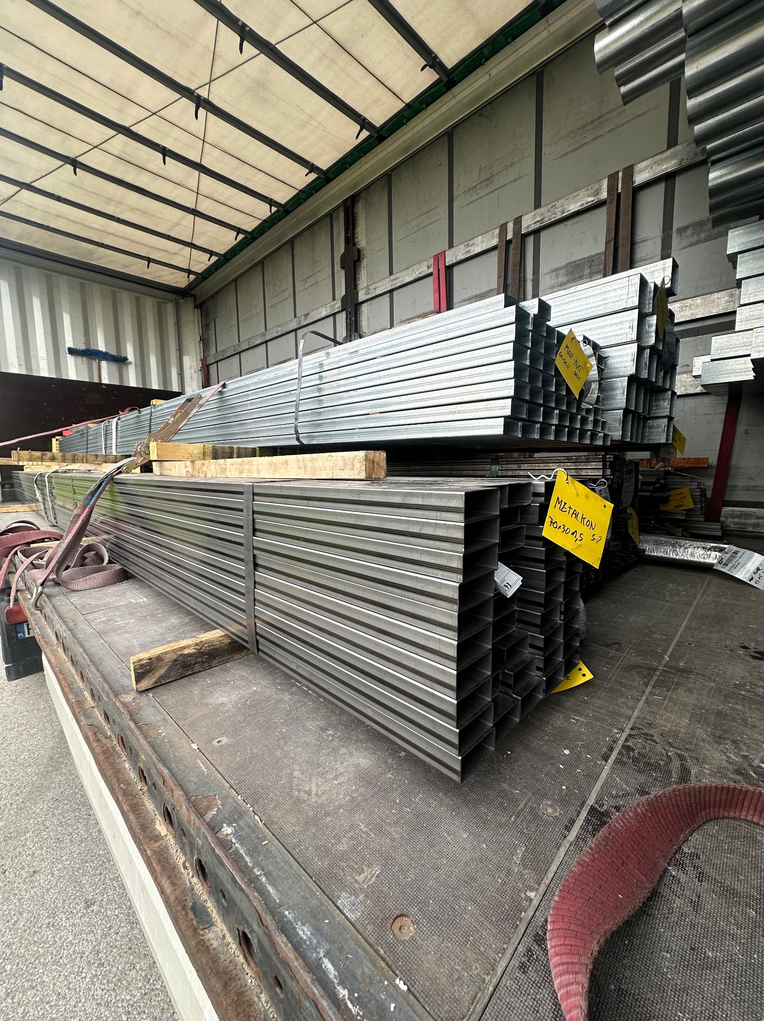 Stalowe profile zamknięte, ułożone w paczki i zabezpieczone pasami transportowymi na platformie ciężarówki. Żółte etykiety z opisem przymocowane do pakietów.