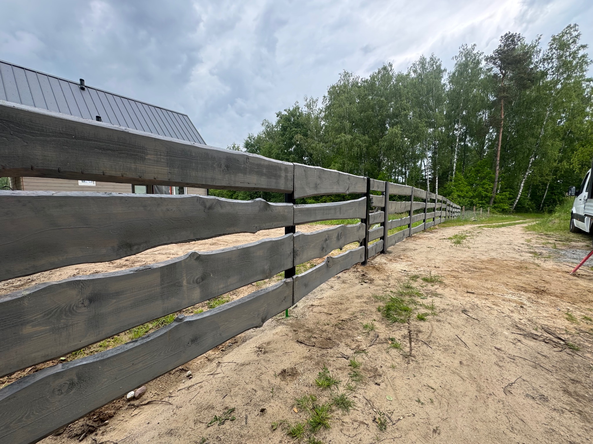 Długi, drewniany płot w kolorze szarości z surowym wykończeniem desek, otaczający posesję. W tle widoczne drzewa i fragment budynku z metalowym dachem.