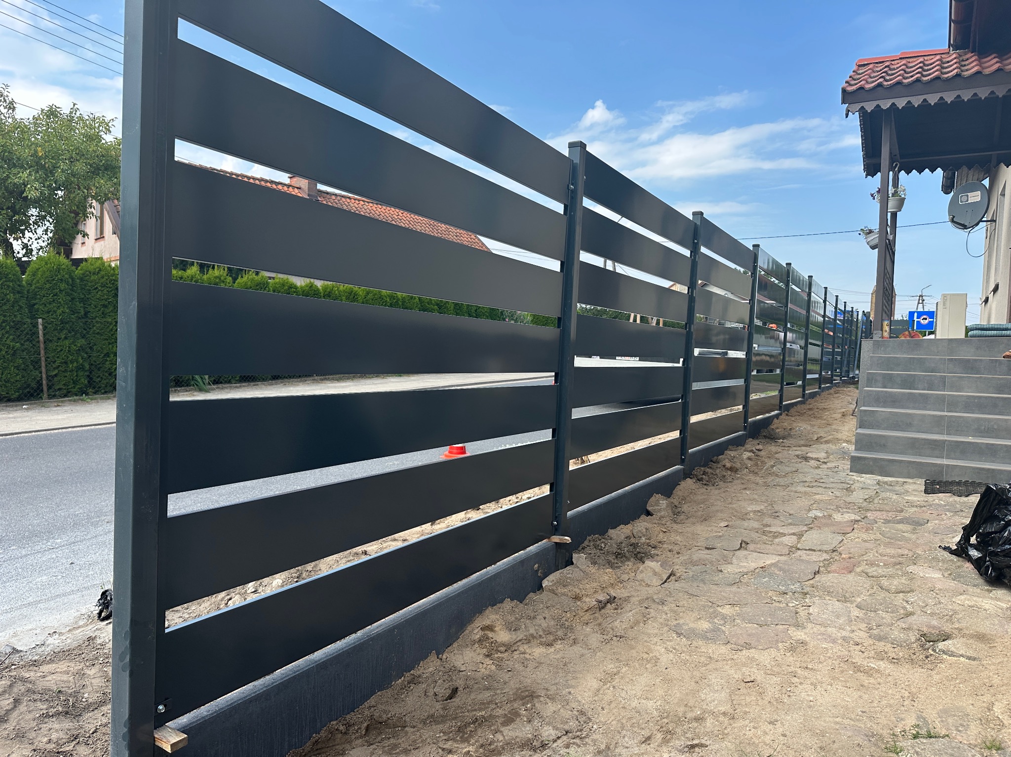 Nowoczesne, poziome ogrodzenie panelowe w kolorze grafitowym, z widocznymi przerwami między panelami, oddzielające posesję od ulicy. W tle dom z elementami drewnianymi.