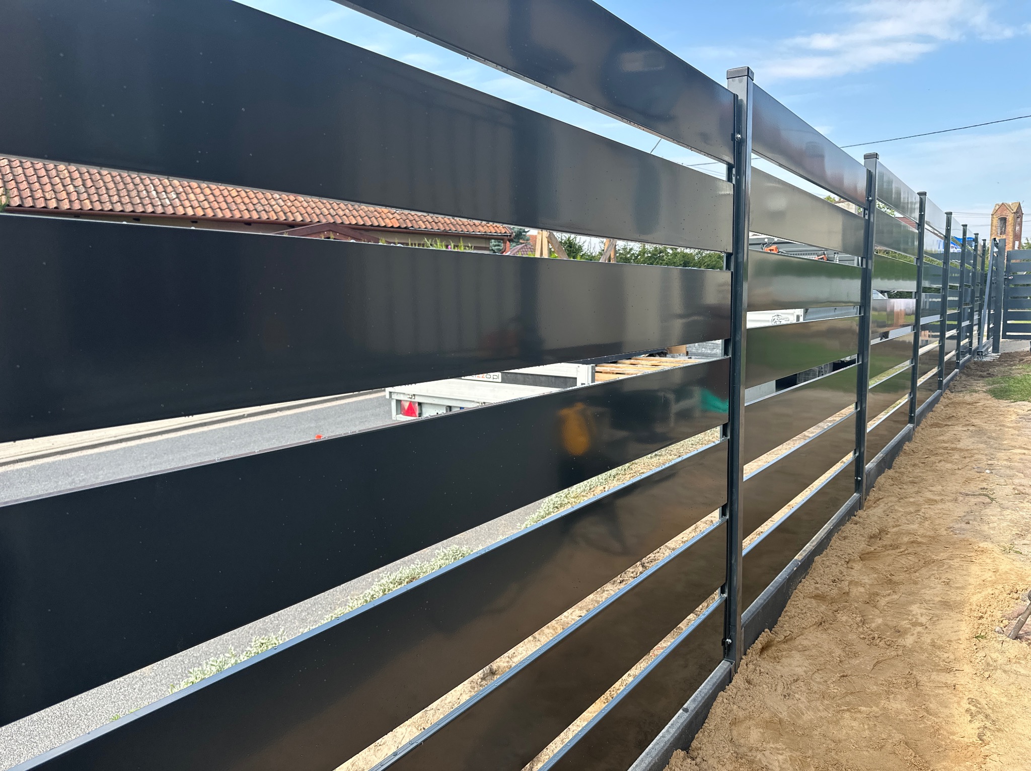 Nowoczesne, czarne ogrodzenie panelowe z poziomymi szczeblami, widok z boku, z fragmentami budynku w tle. Świeżo zamontowane, z widoczną ziemią przy podstawie.