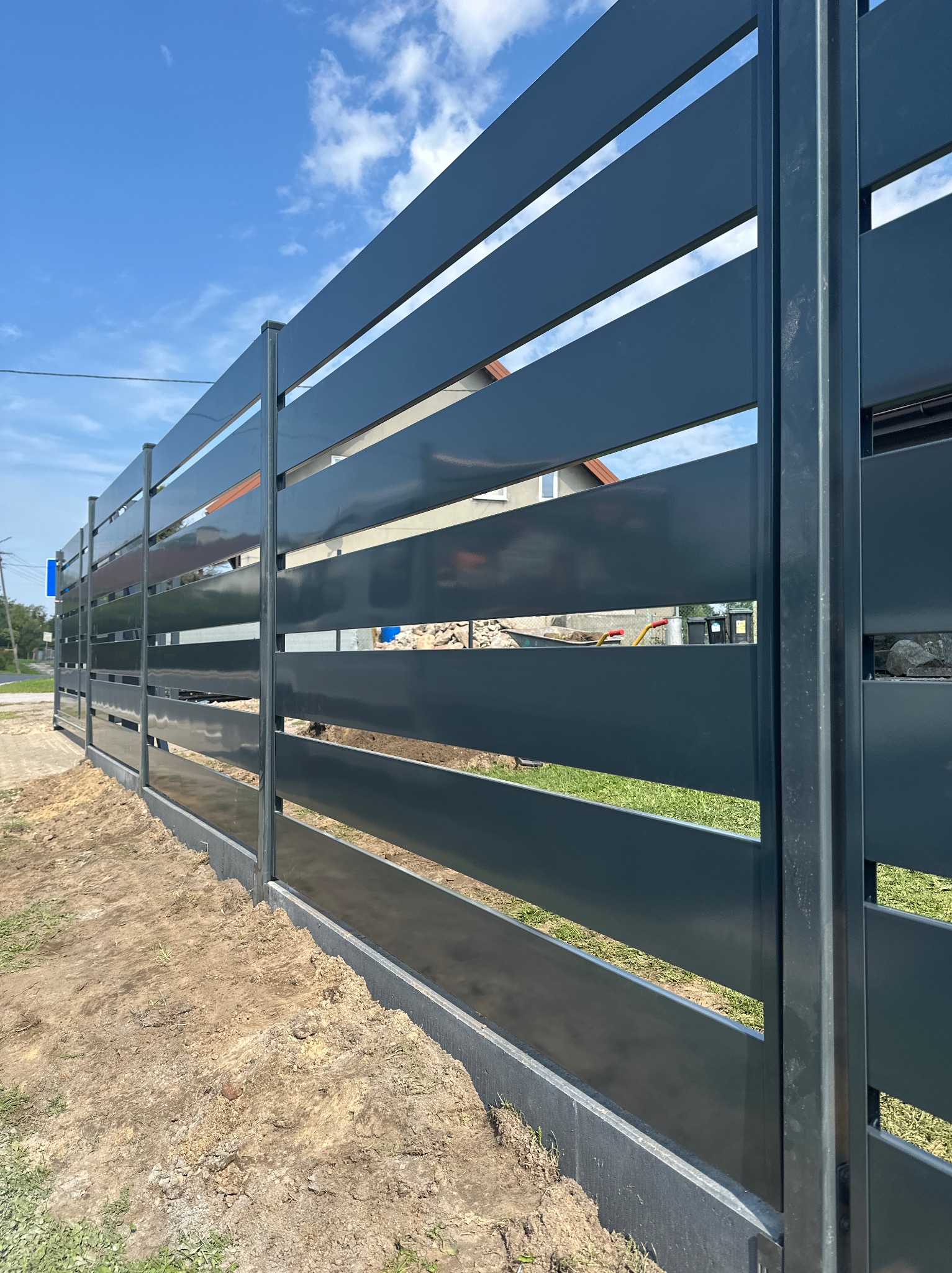 Nowoczesne, poziome ogrodzenie panelowe w kolorze grafitowym, z widocznymi prześwitami między panelami. W tle dom jednorodzinny pod błękitnym niebem.