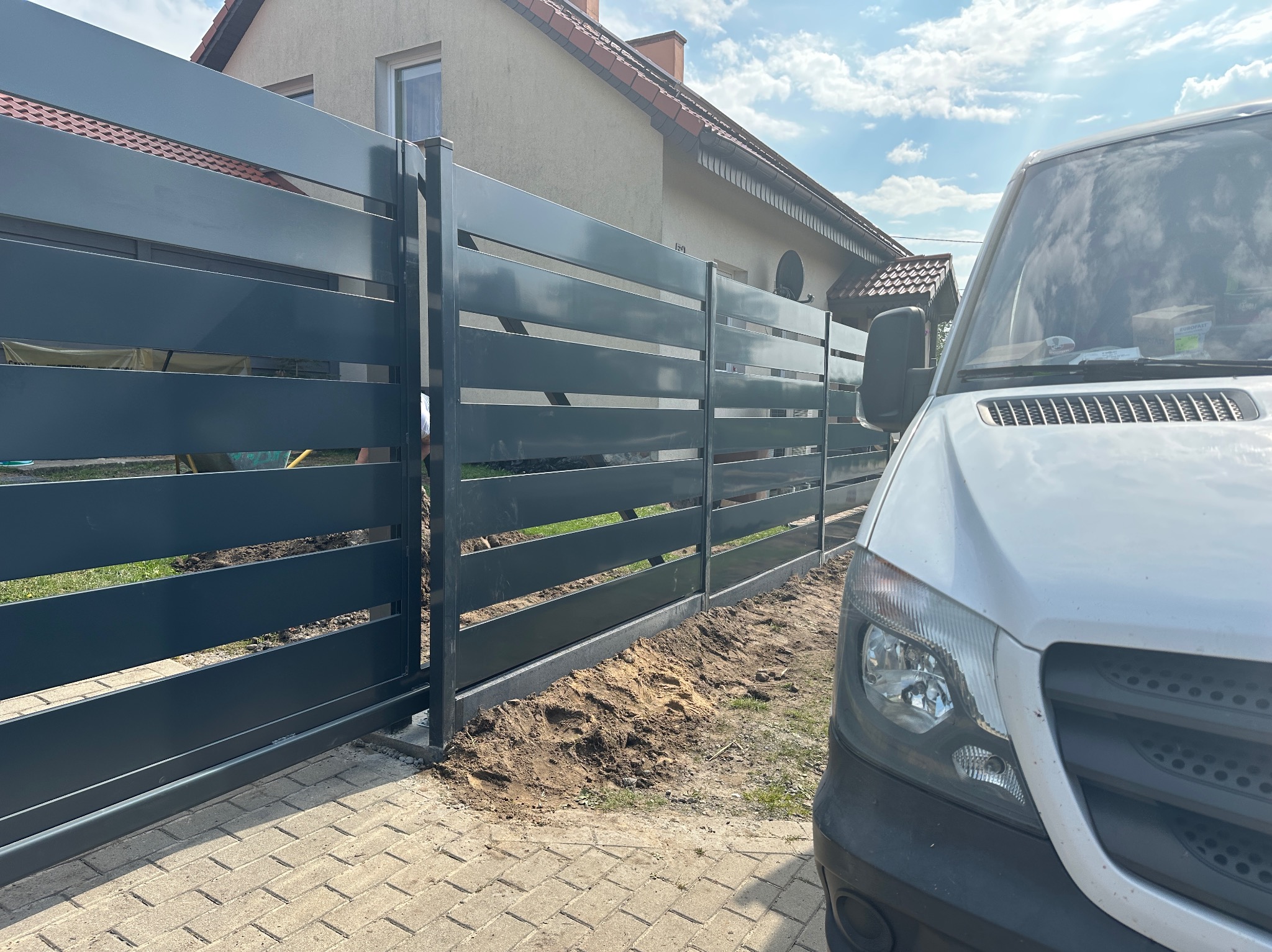 Nowoczesna, przesuwna brama panelowa w kolorze grafitowym, widoczna z perspektywy ulicy, z białym samochodem dostawczym po prawej stronie. Dom jednorodzinny w tle.