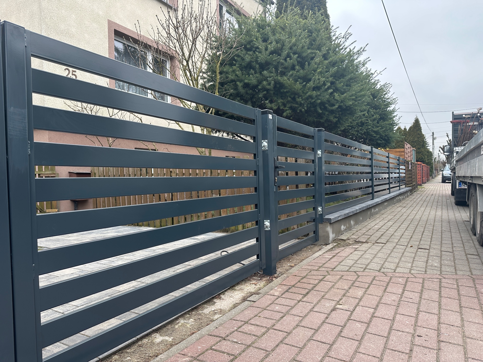 Grafitowe ogrodzenie posesji z poziomych paneli, numer domu 25, widok z zewnątrz na tle zieleni i kostki brukowej. Nowoczesny design, poziome ułożenie paneli.