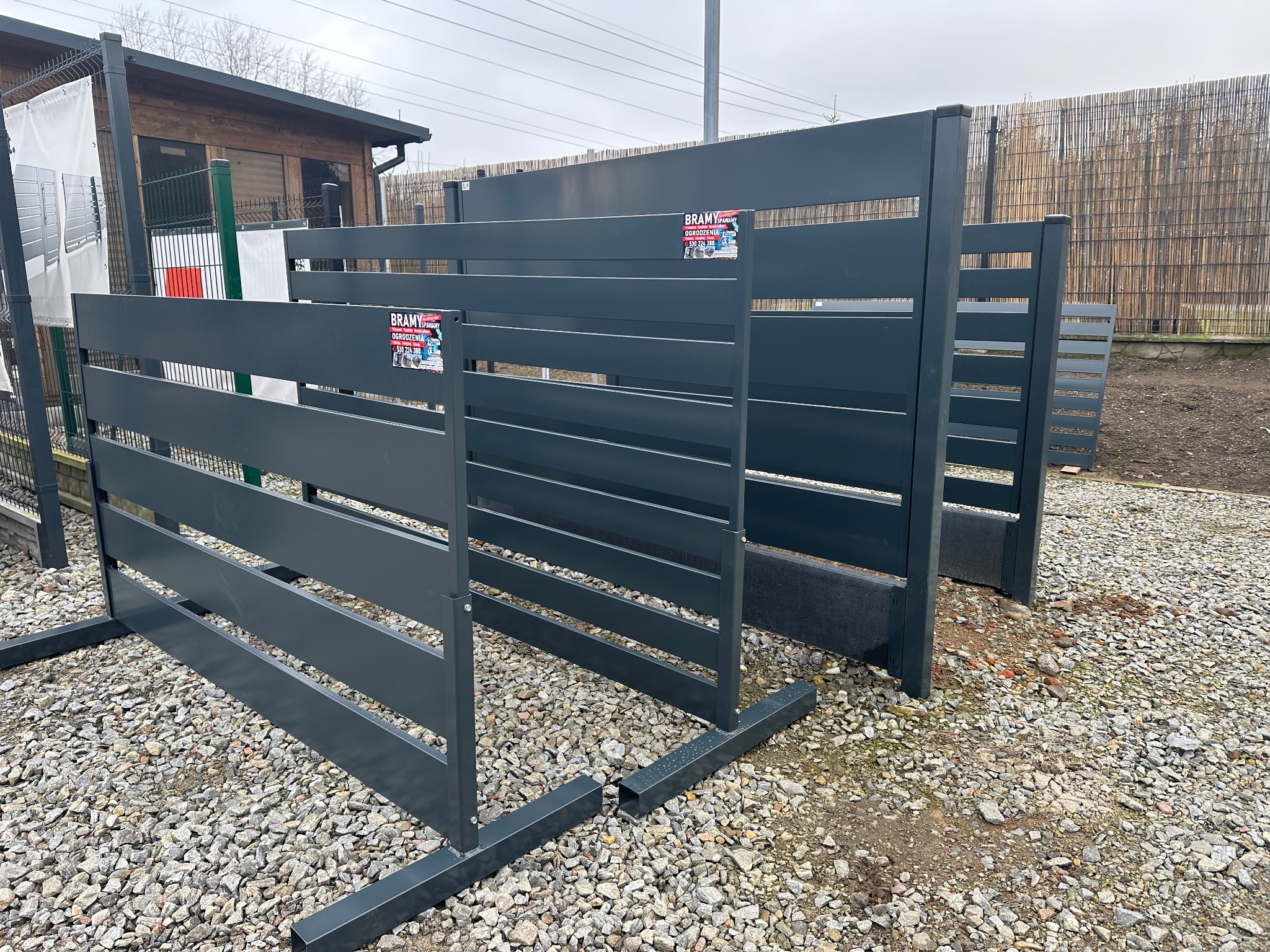 Nowoczesne, grafitowe panele ogrodzeniowe z poziomymi szczeblami, prezentowane na żwirowym podłożu. Widoczne detale montażowe i naklejka producenta.