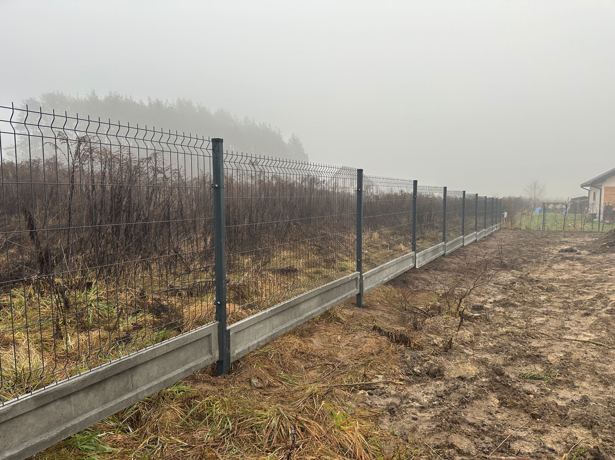 Szary panelowy płot z podmurówką na zewnątrz, w tle zalesiony teren spowity mgłą. Widoczne słupki i siatka ogrodzeniowa. Teren wokół ogrodzenia jest nierówny i błotnisty.
