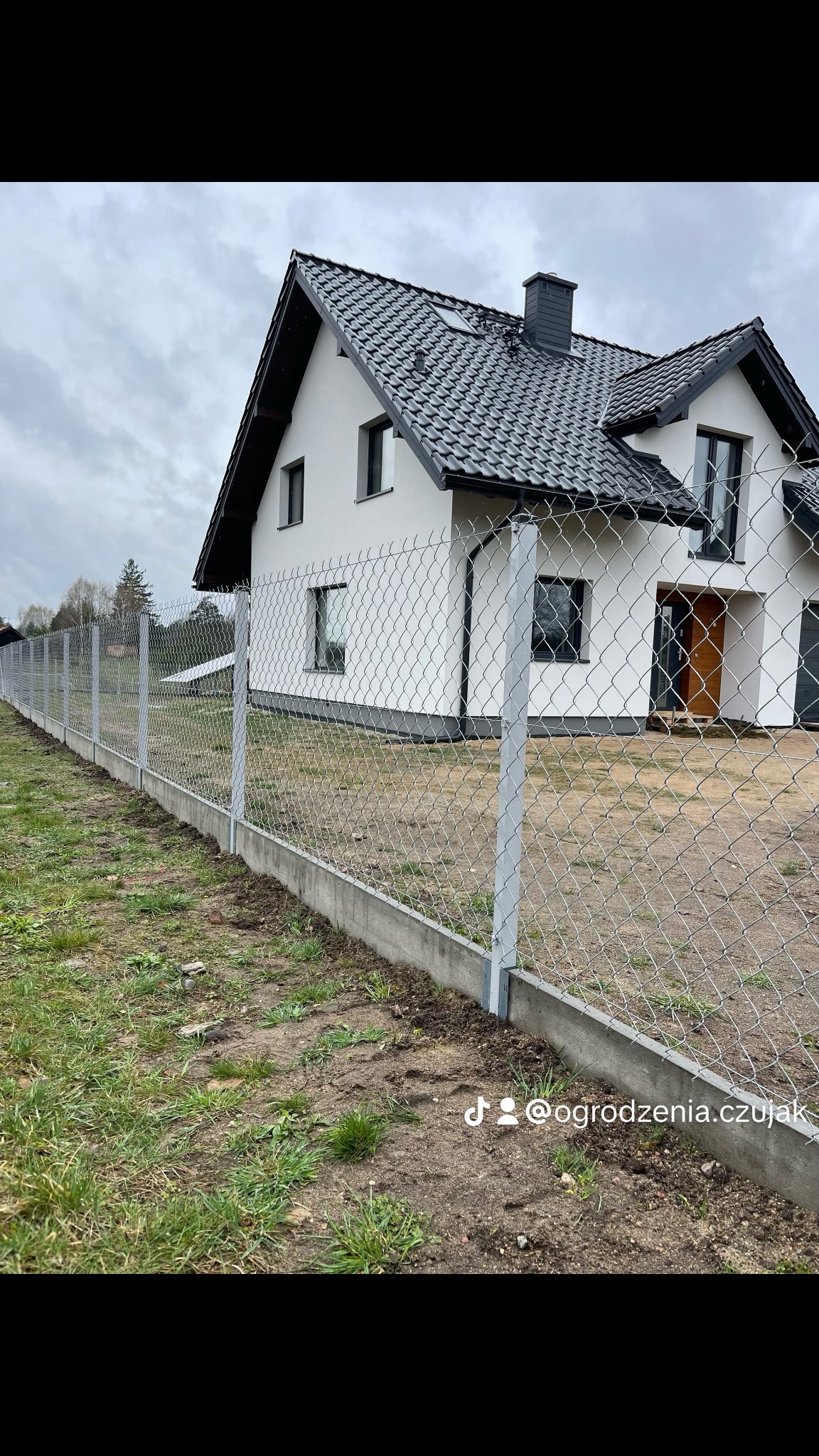 Długa siatka ogrodzeniowa z betonową podmurówką, otaczająca posesję z nowoczesnym domem. Szara, pochmurna pogoda. Widoczne logo firmy na dole zdjęcia.