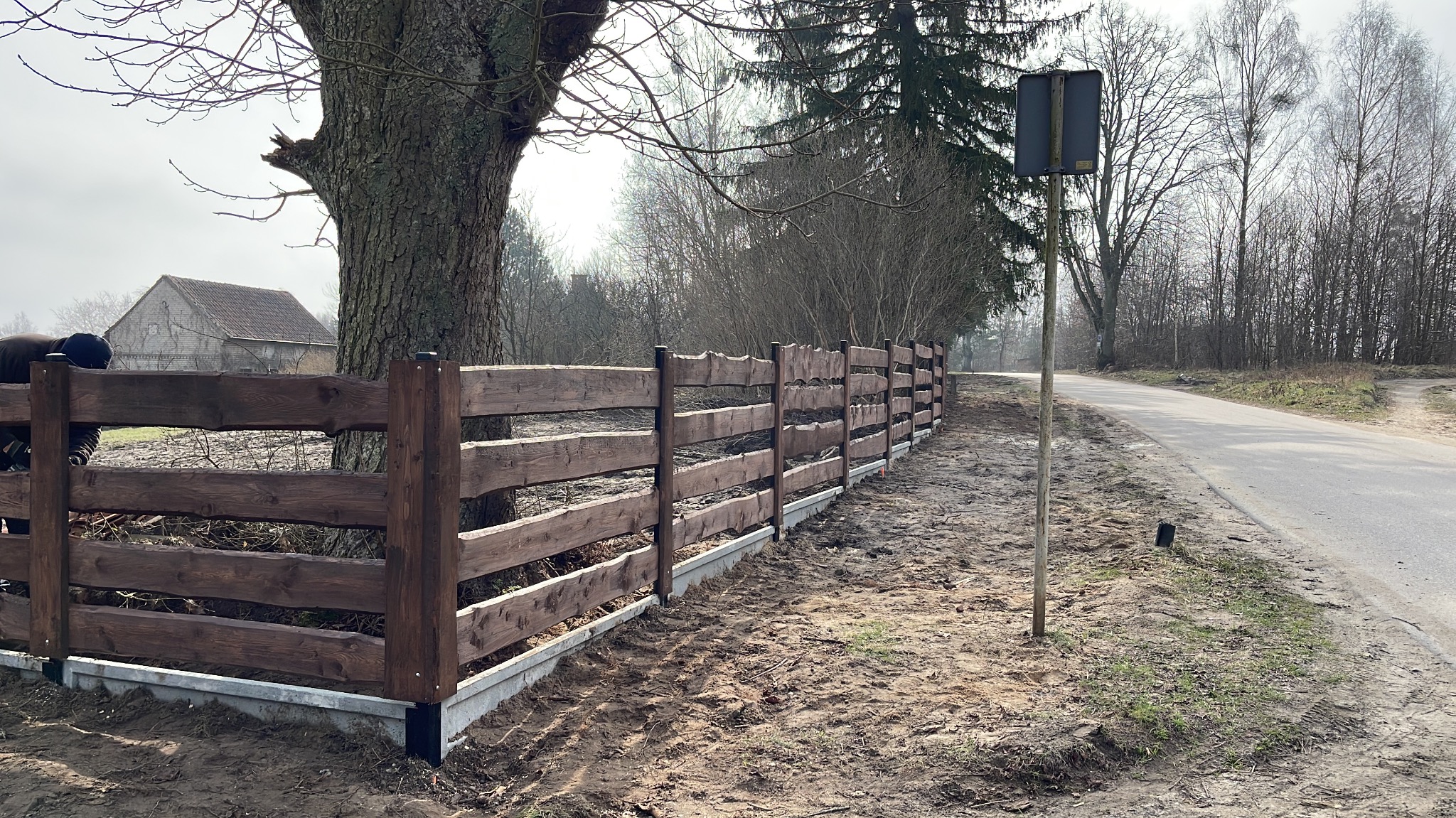 Drewniane ogrodzenie posesji ze słupkami, Szczytno. Widok z boku na długi płot z brązowych desek, betonowa podmurówka. W tle drzewa i budynek.