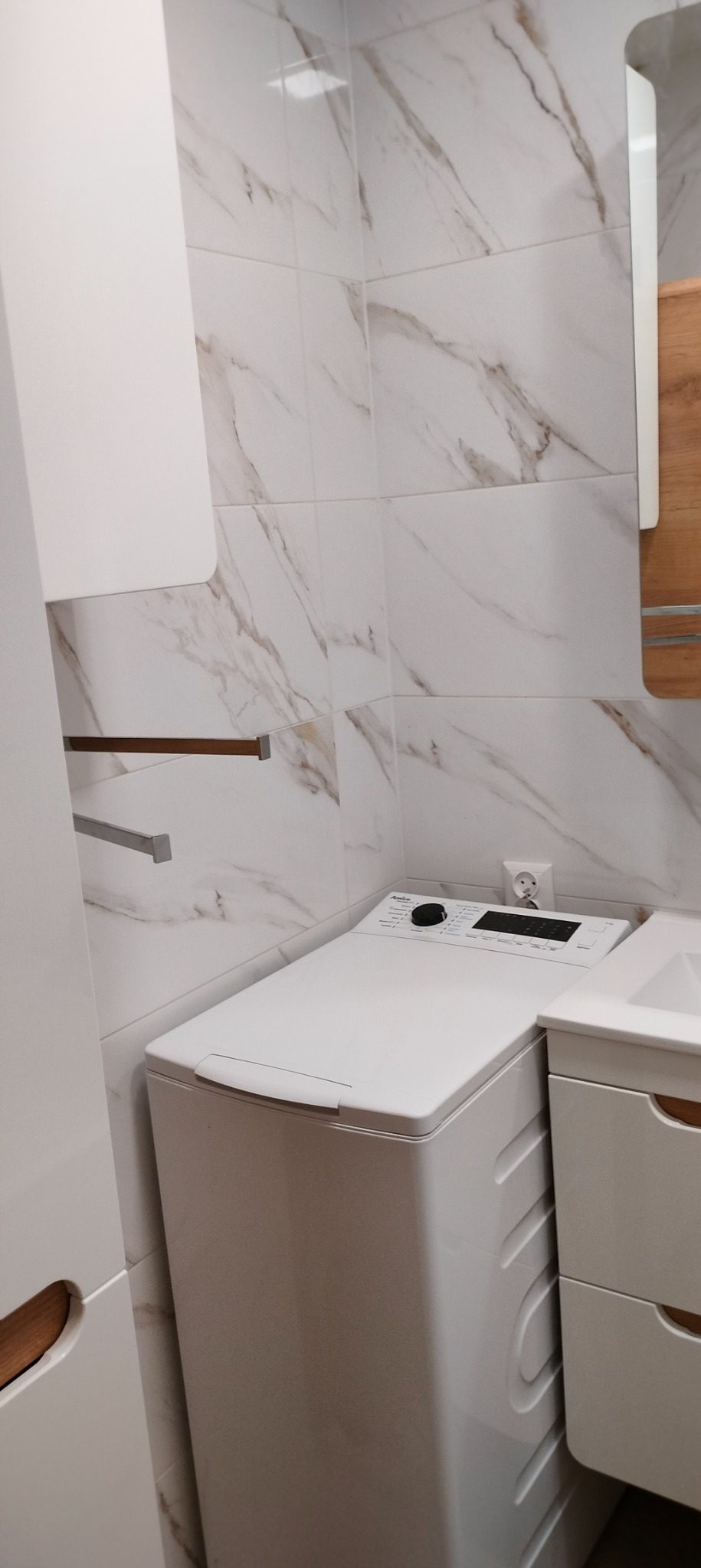 Wykończona łazienka w Łodzi: białe marmurowe płytki ścienne, pralka marki Amica zintegrowana z szafką umywalkową, widoczne gniazdko elektryczne.