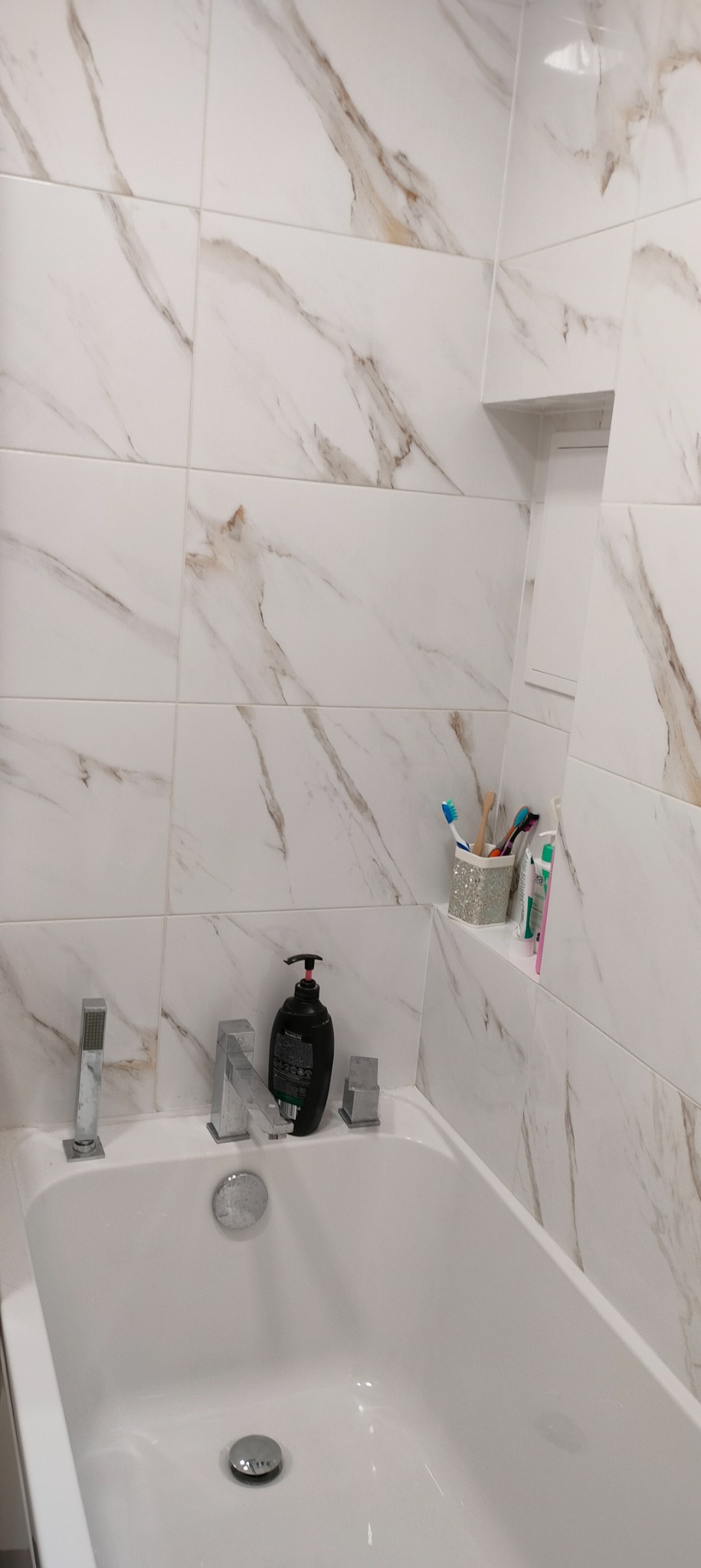 Biała wanna z nowoczesną armaturą i marmurowymi płytkami ściennymi, wnęka z akcesoriami łazienkowymi.