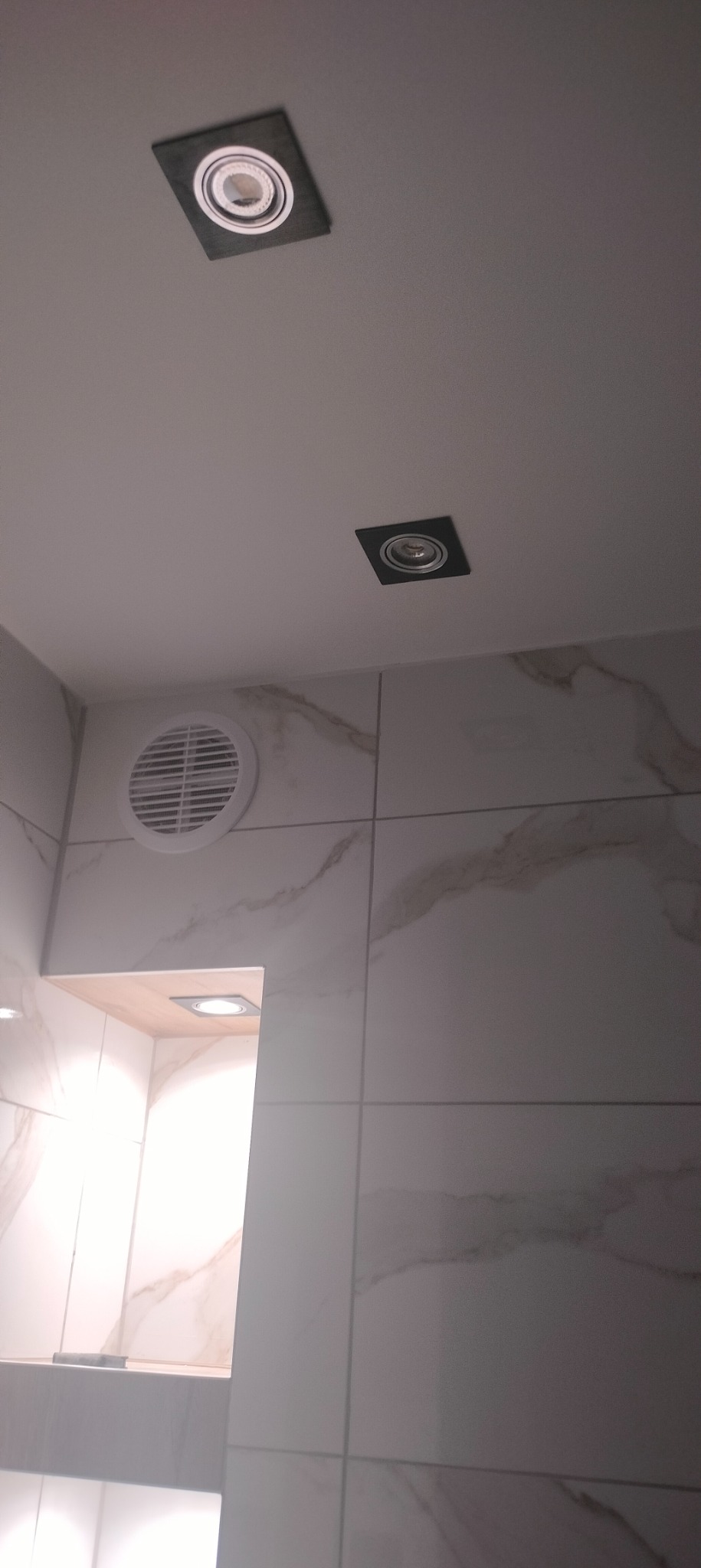 Dwie kwadratowe lampy sufitowe typu spot wpuszczone w biały sufit, widoczny fragment ściany z białych płytek imitujących marmur oraz okrągły wywiewnik wentylacyjny.