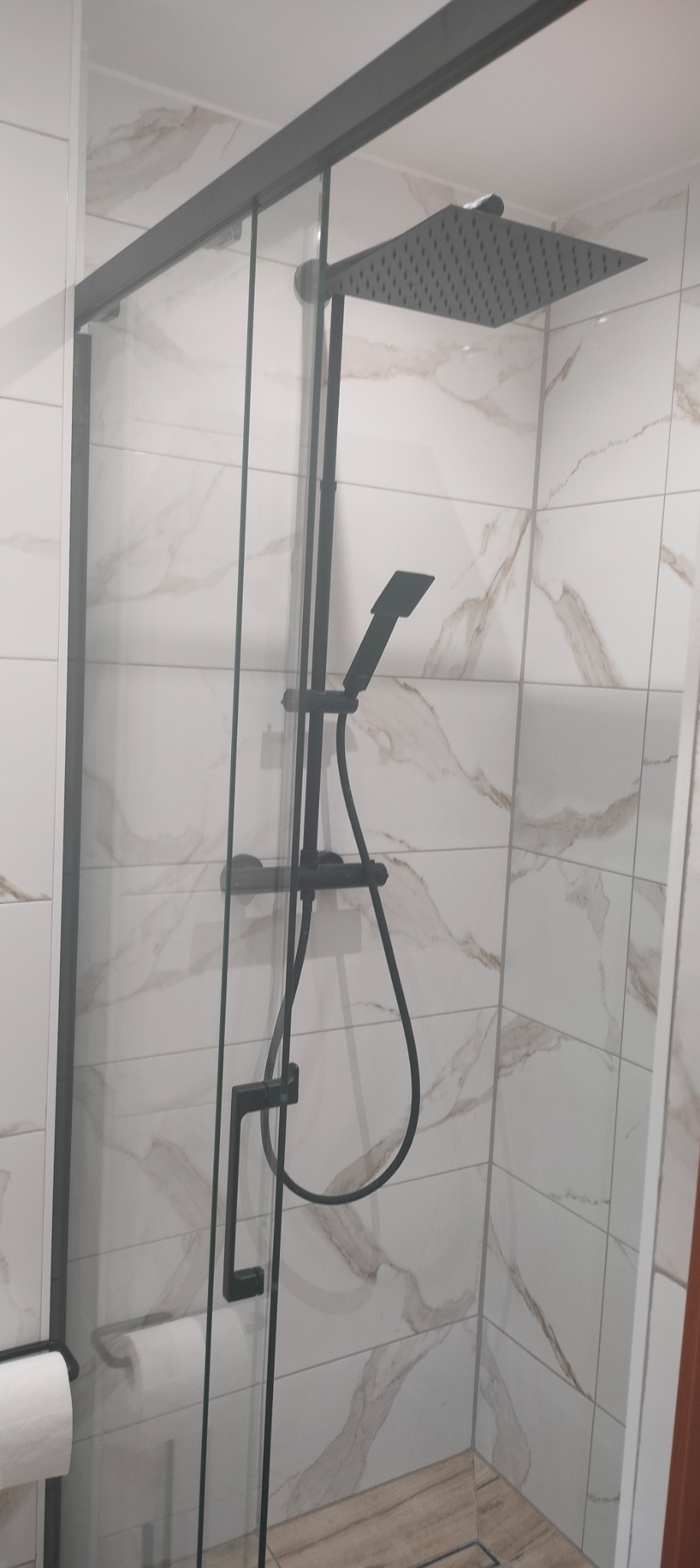 Nowoczesna kabina prysznicowa z czarną armaturą, deszczownicą i słuchawką, wyłożona płytkami imitującymi marmur.